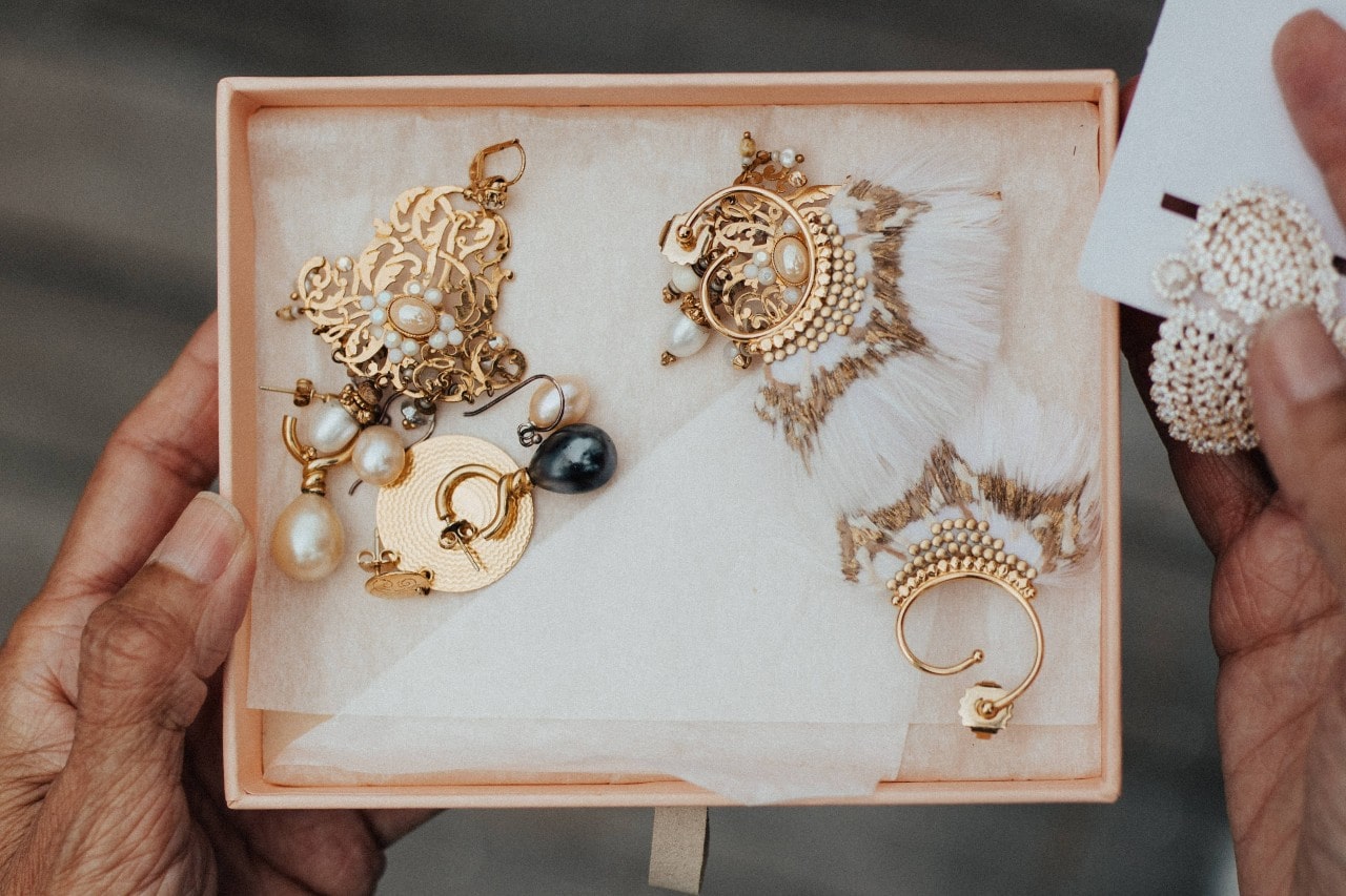A pair of hands holds a jewelry box filled with ornate gold earrings, featuring pearls, filigree patterns, and intricate designs.