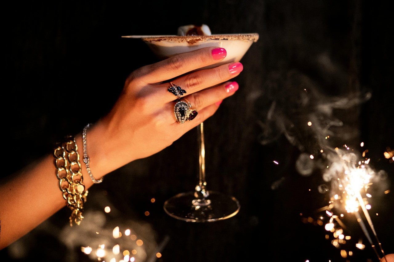 A woman's hand with bright pink nails holds a cocktail glass, adorned with bold statement rings and gold bracelets amid festive sparklers.