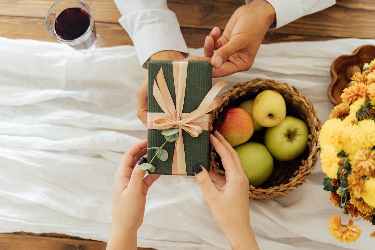 Two hands exchange a green wrapped gift with a peach satin ribbon, placed on a table decorated with apples, flowers, and a glass of red wine.