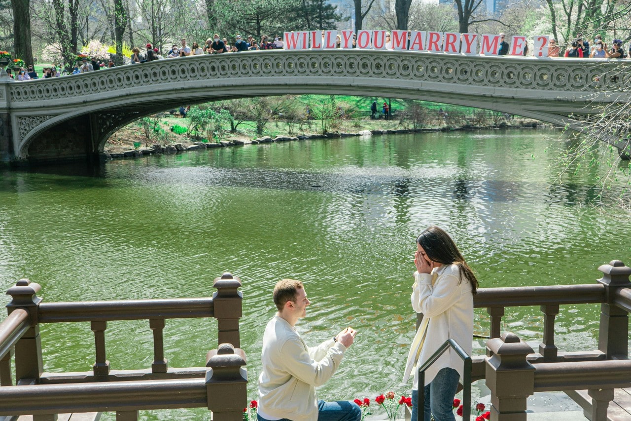 A close up of a man standing on his knee proposing to a woman in front of the river and bridge.