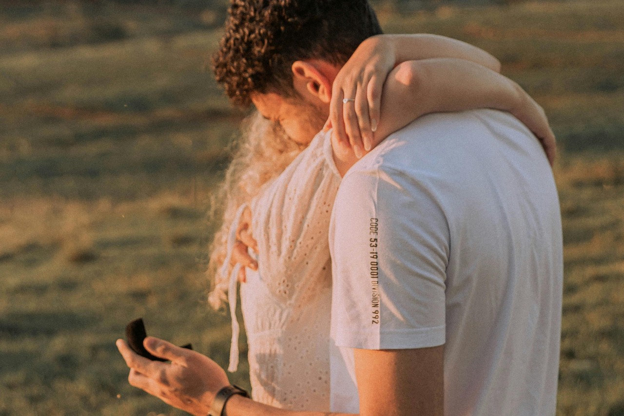 A man in a white t-shirt embracing a woman in a white embroidered top, while holding an engagement ring box in one hand.