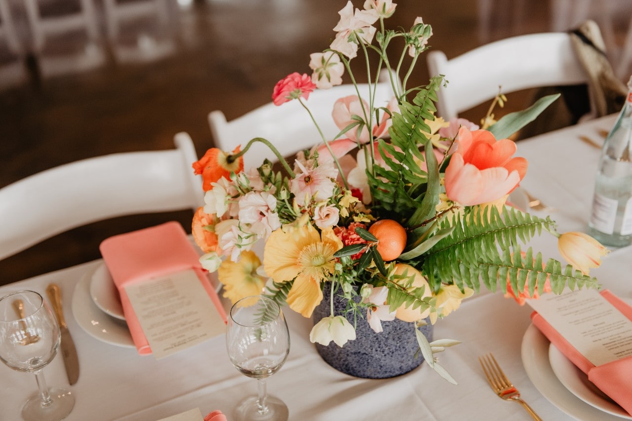 A colorful floral centerpiece with coral napkins and gold flatware decorates an elegantly set wedding reception table