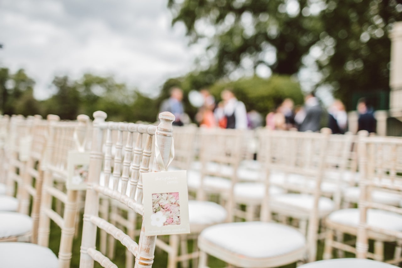 Rows of white chairs are arranged for an outdoor wedding ceremony, with guests gathered in the softly blurred background.