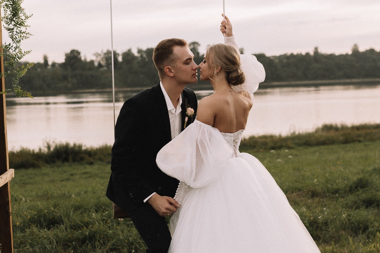 A bride and groom share a kiss while sitting on a swing beside a calm lakeside landscape