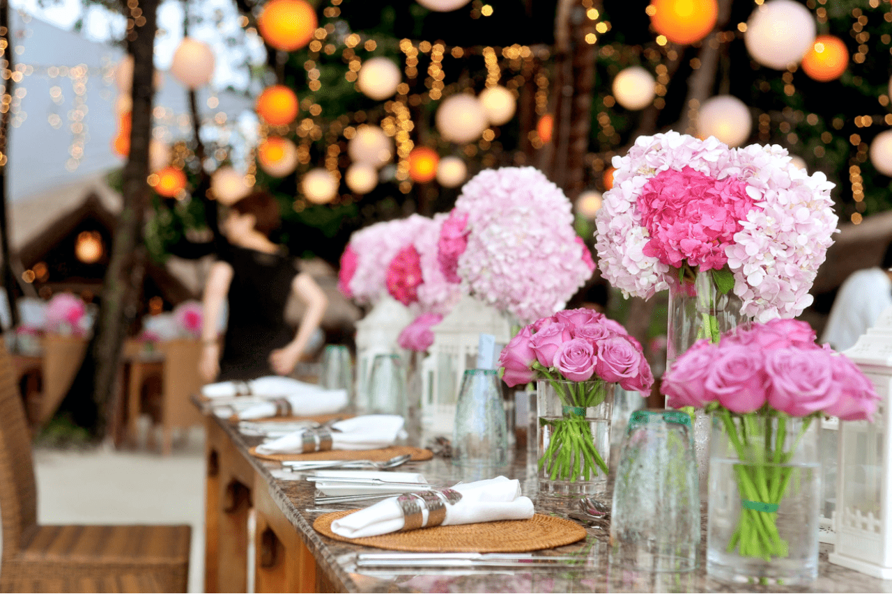 A beautifully styled outdoor reception table is decorated with pink floral centerpieces and glowing string lights for a romantic wedding setting.