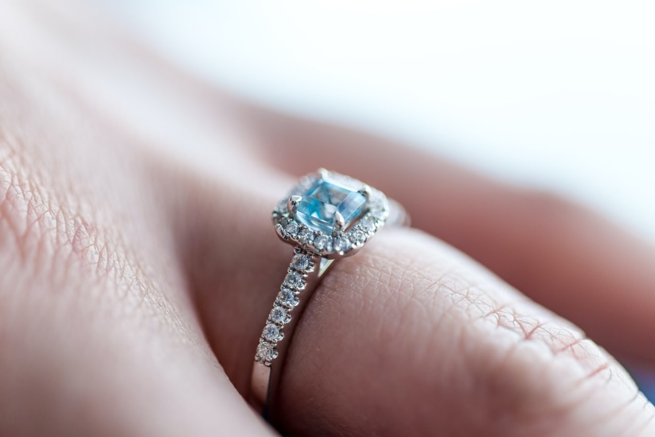 A close-up of a blue engagement ring with a diamond halo on a person’s finger.