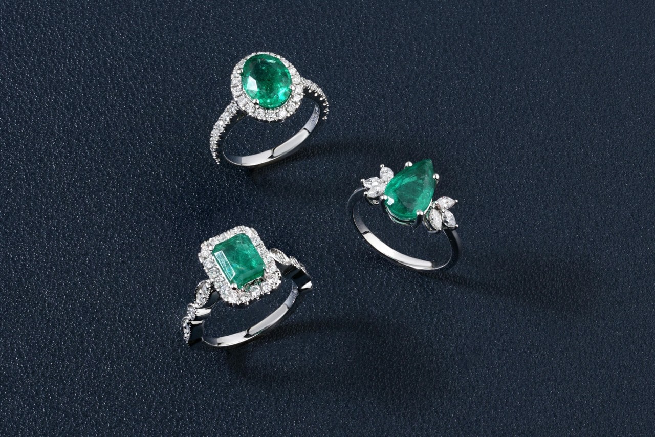 Three emerald engagement rings of different cuts, sitting on a navy surface.