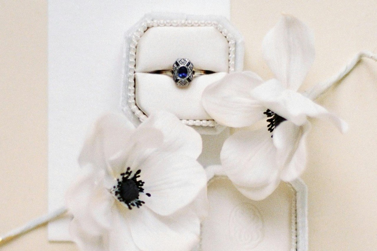 A stunning engagement ring in a white ring box with a pearl framing and some white flowers surrounding it.