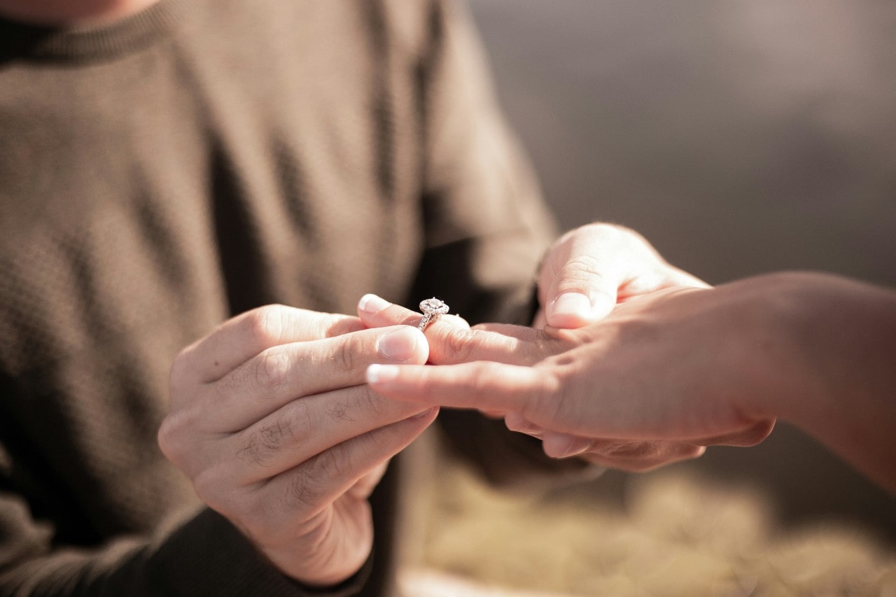 A man sliding an engagement ring onto a woman&rsquo;s hand.
