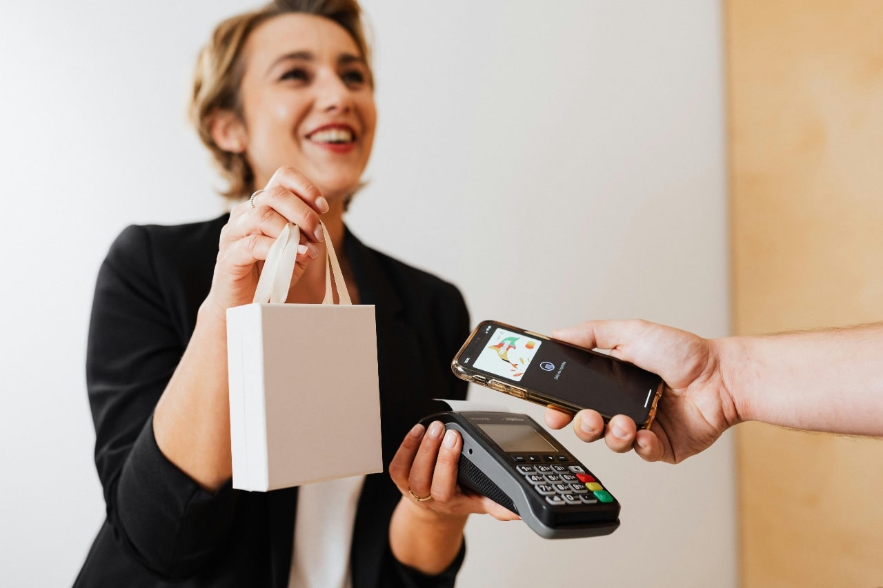 A smiling woman holds a small shopping bag as a person makes a payment using a smartphone at a card reader