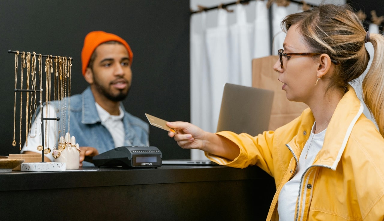 A woman in a yellow jacket hands a credit card to a cashier in an orange beanie and denim jacket, with gold jewelry displayed on the counter.