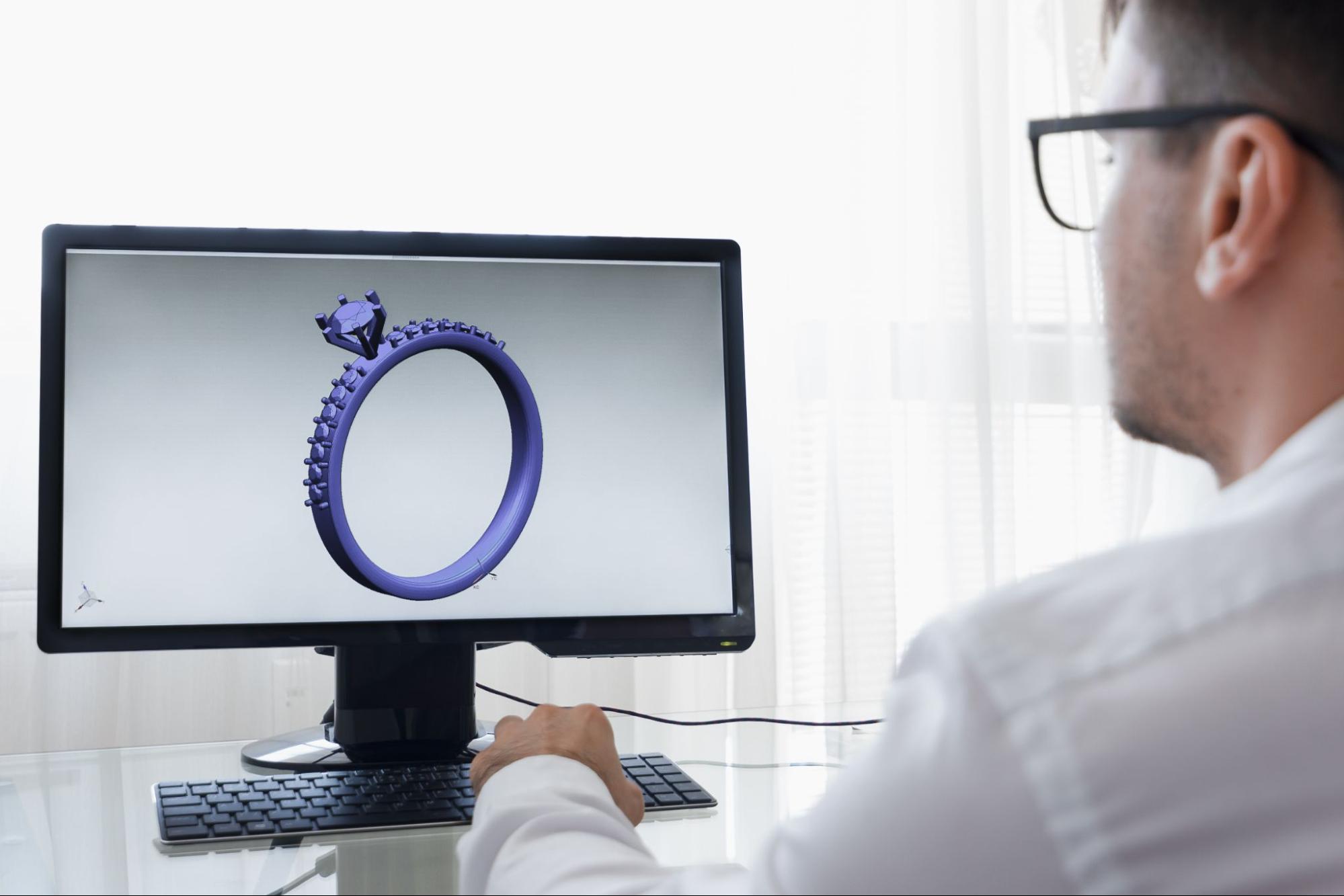 A man views a 3D digital model of a diamond engagement ring on a computer screen, using CAD software for jewelry design.
