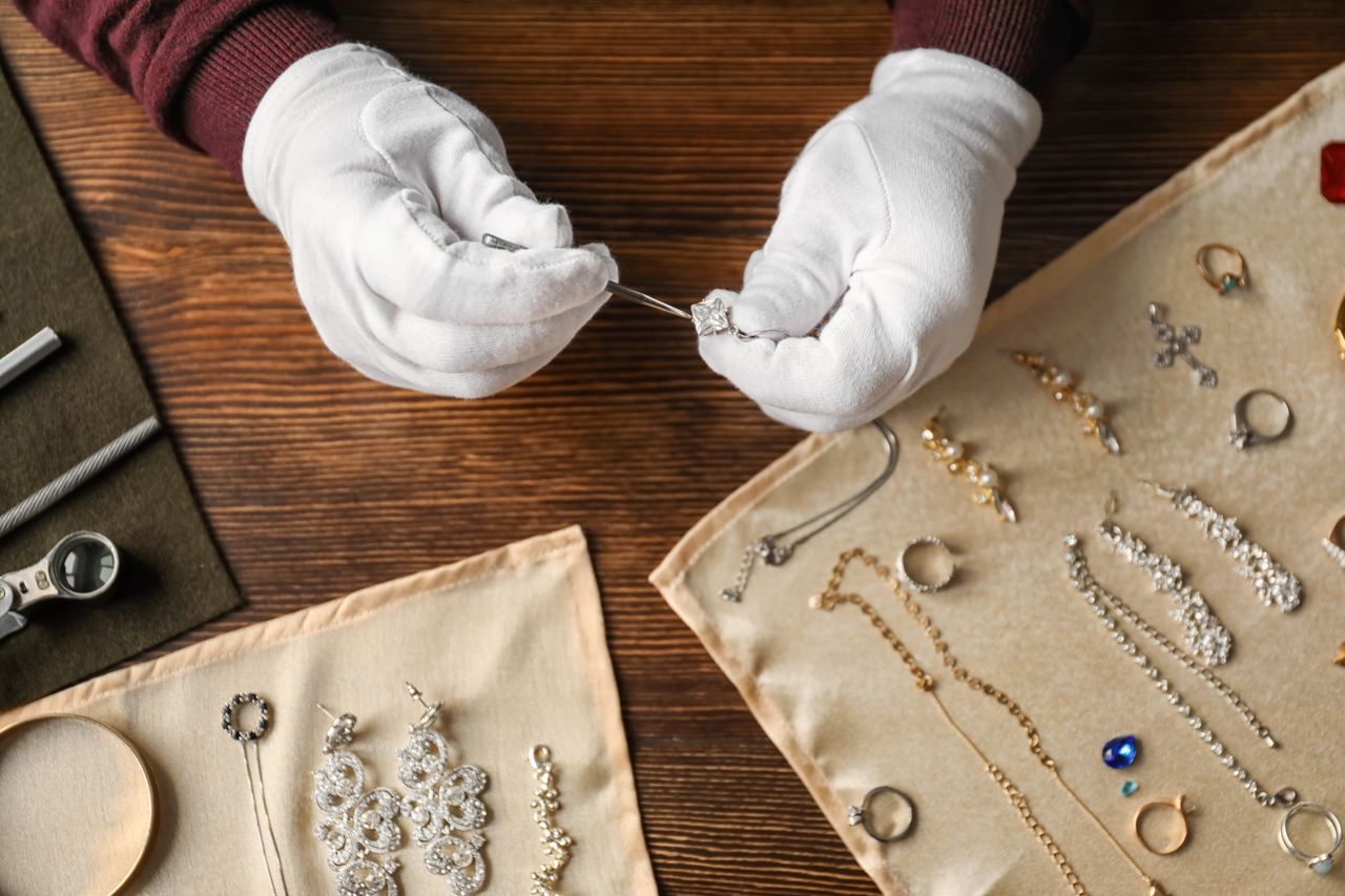 A close up of a jeweler in white gloves uses tools to examine a diamond ring on a wooden table surrounded by various designs of jewelry and repair instruments.