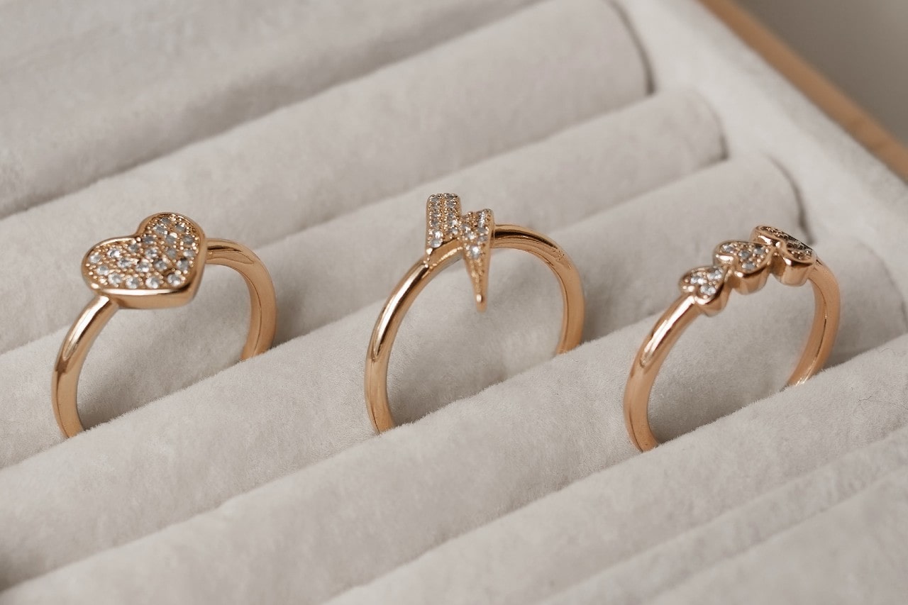 A close up of 3 yellow gold fashion rings with sparkling diamonds with different center shapes resting in individual slots within a light-colored jewelry display.