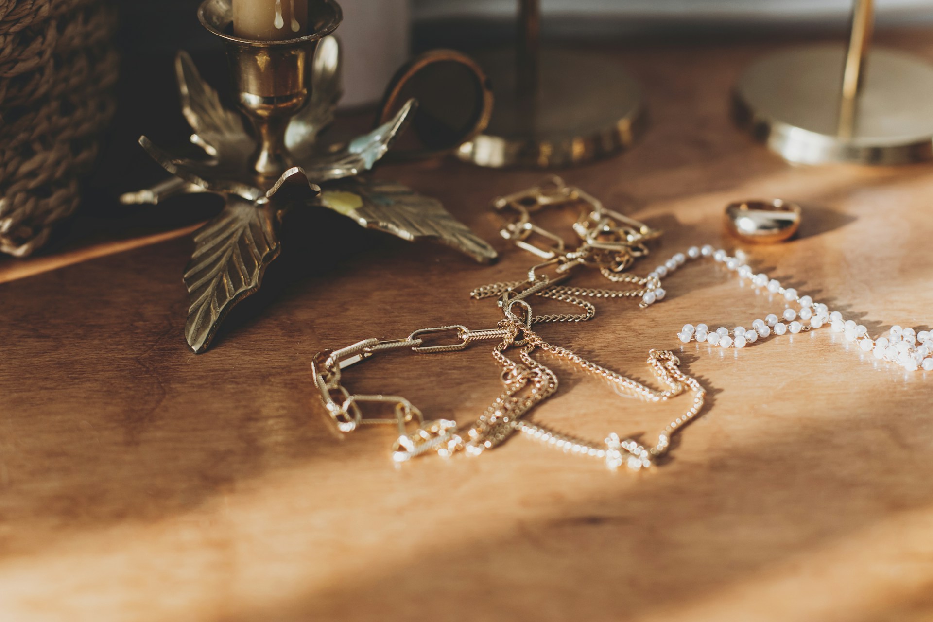 A close-up of gold chains, pearl necklaces, and a ring laid out on a wooden surface near brass candle holders.