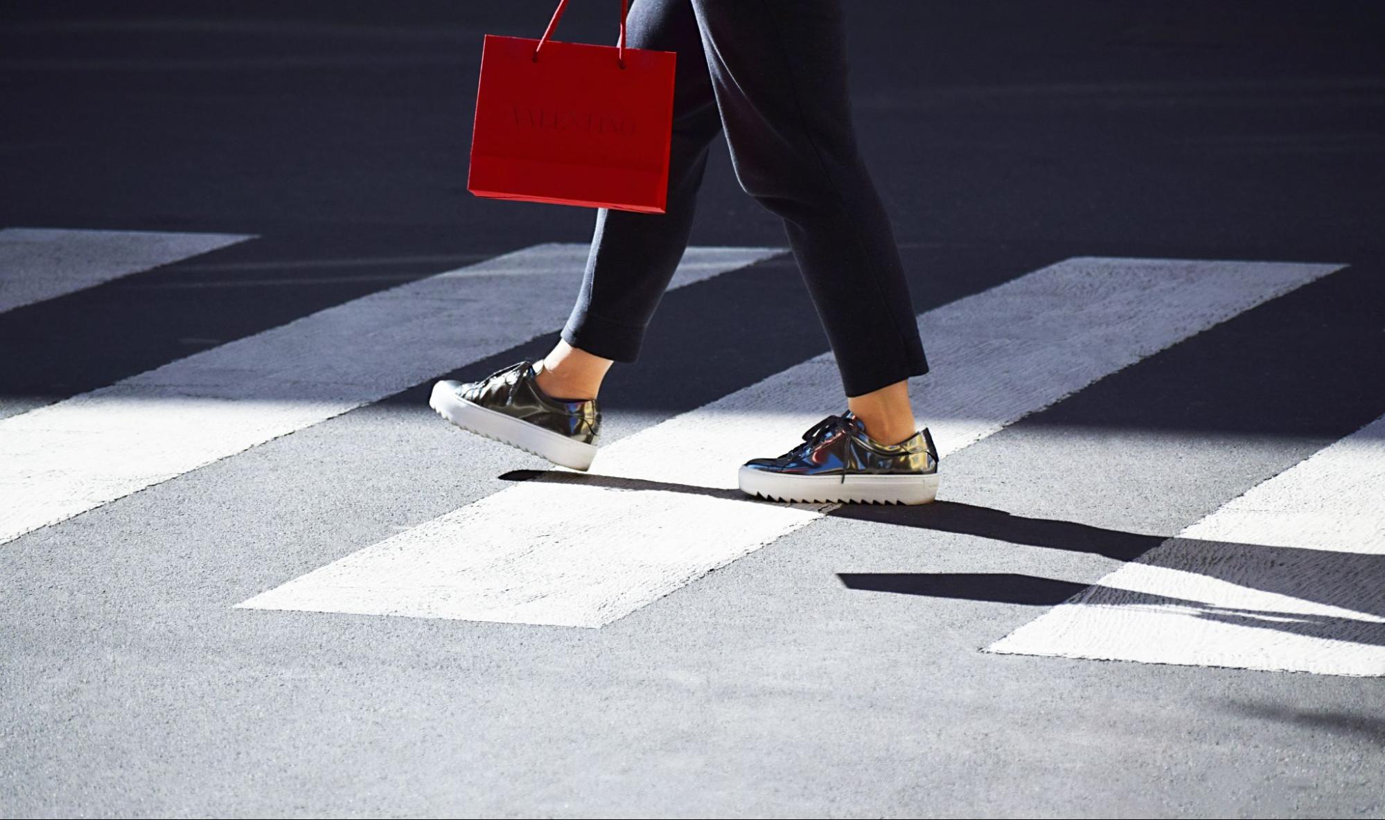 A person wearing shiny silver sneakers and dark pants walks across a sunny crosswalk, holding a red shopping bag.