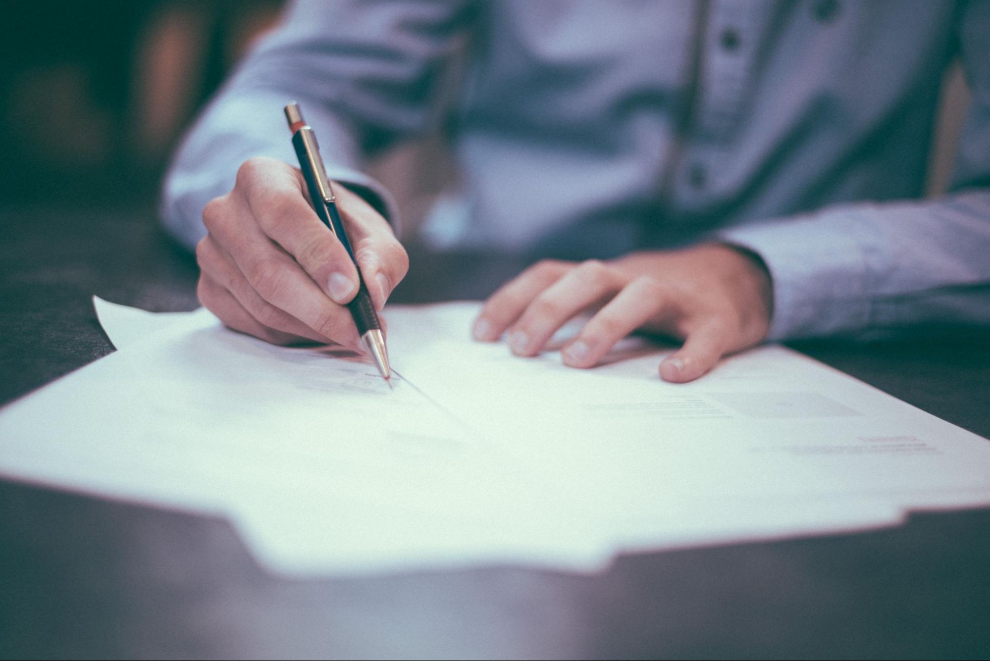 A person in a blue shirt writes on papers with a pen at a desk.