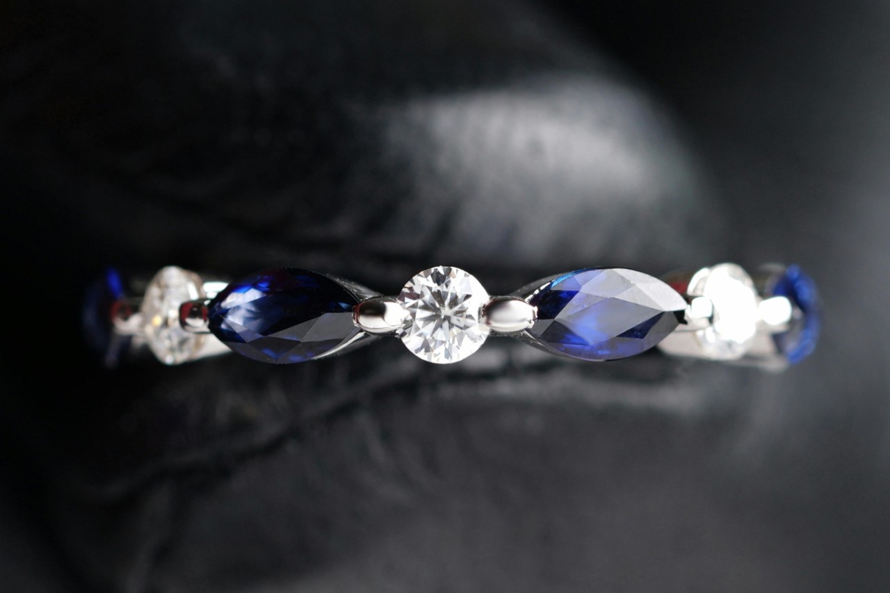 A close-up of a white gold ring featuring alternating round white diamonds and marquise-cut deep blue sapphires
