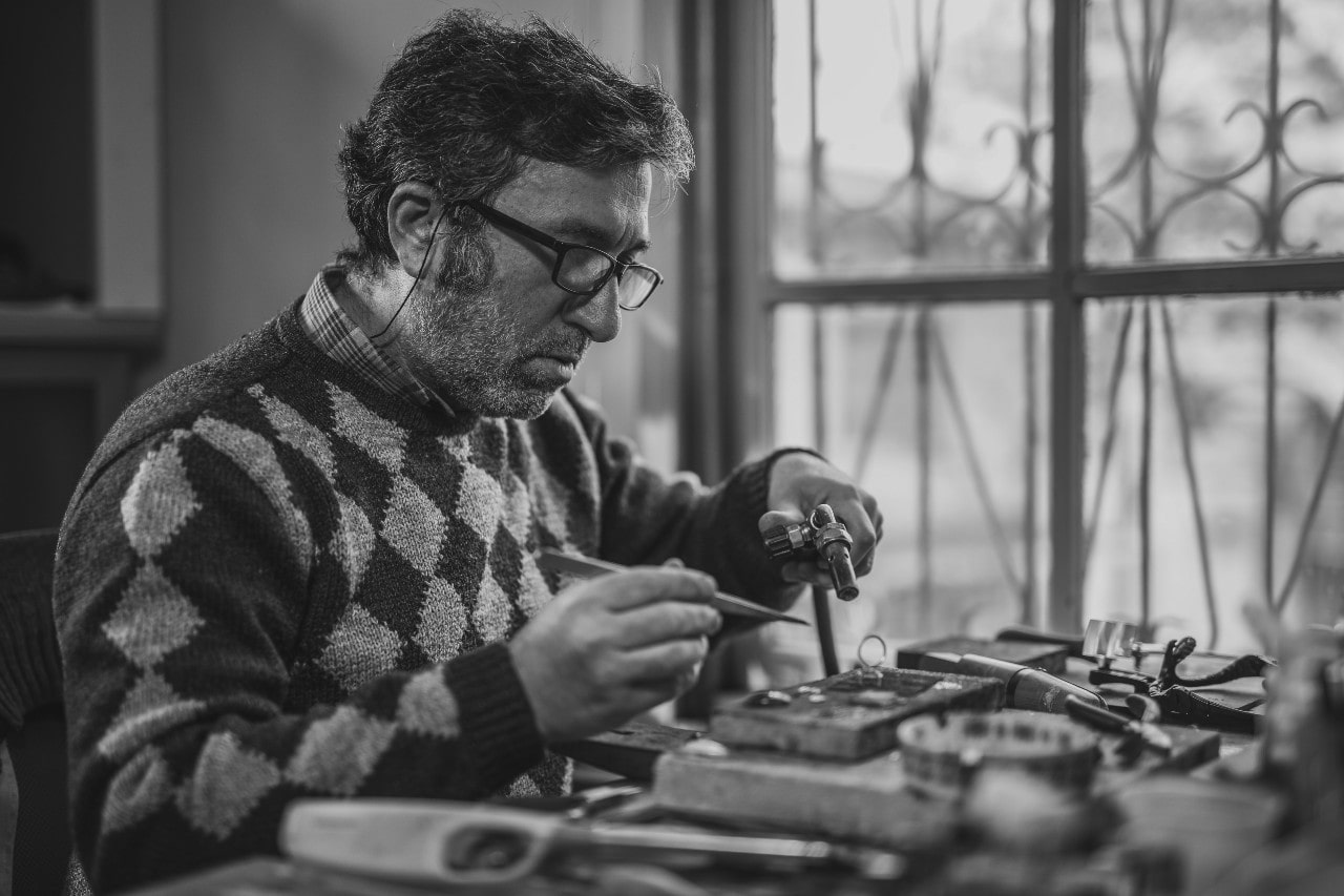 Black-and-white photo of a jeweler in glasses and a sweater repairing jewelry at a cluttered workbench with a window in the background.