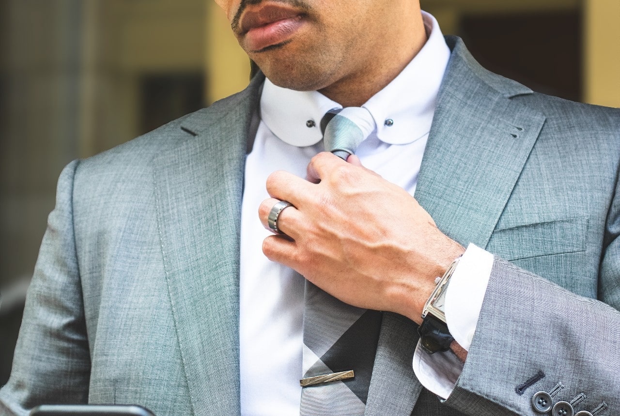 A man in a grey suit fixing his tie, showcasing a titanium wedding band and leather wristwatch.