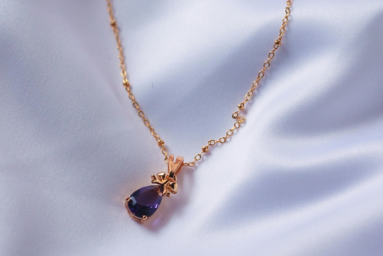 A close-up of a pear-cut purple amethyst pendant necklace on white satin fabric.