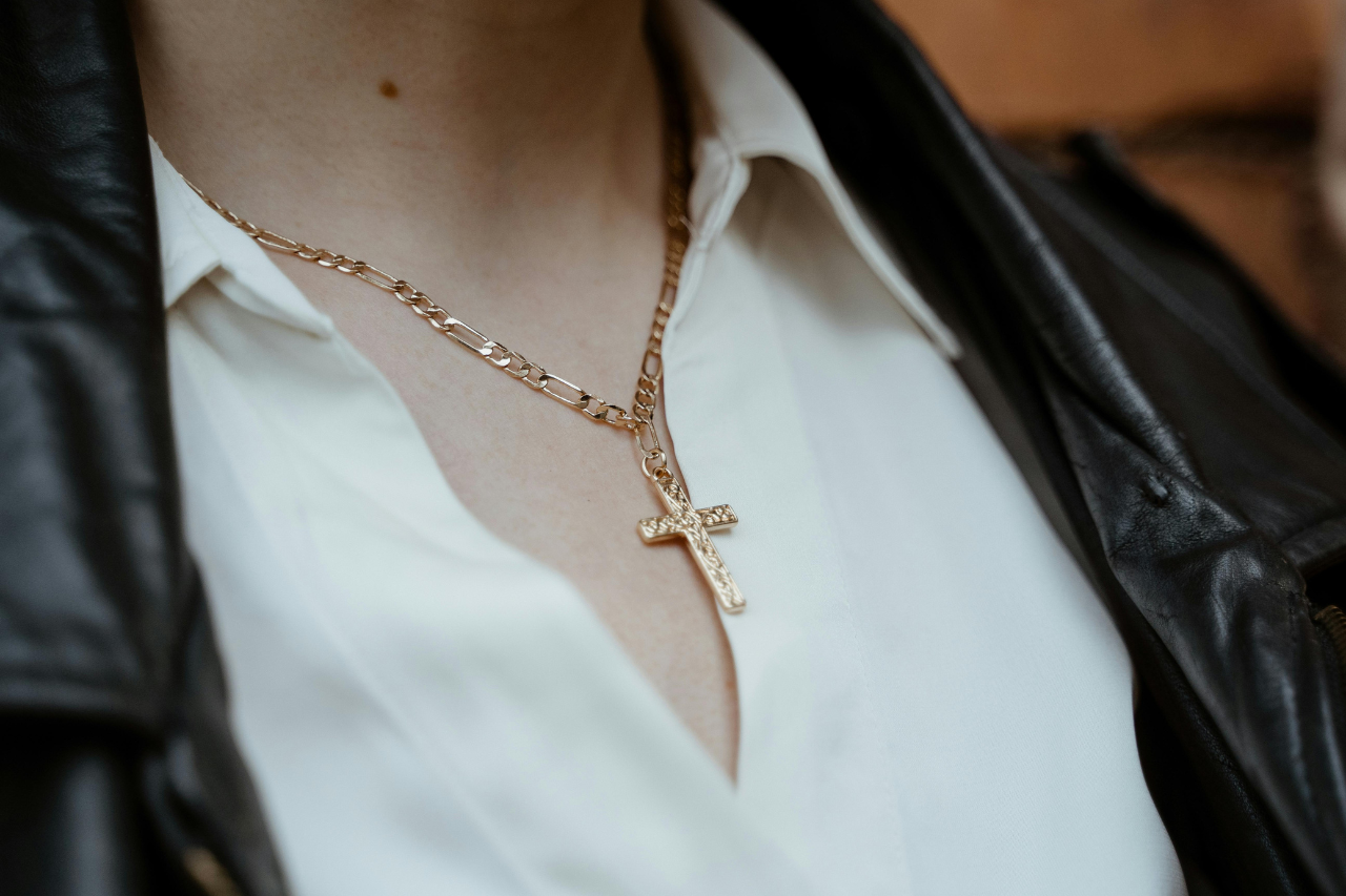 A close-up of a gold figaro chain cross necklace worn with a white shirt and black leather jacket.