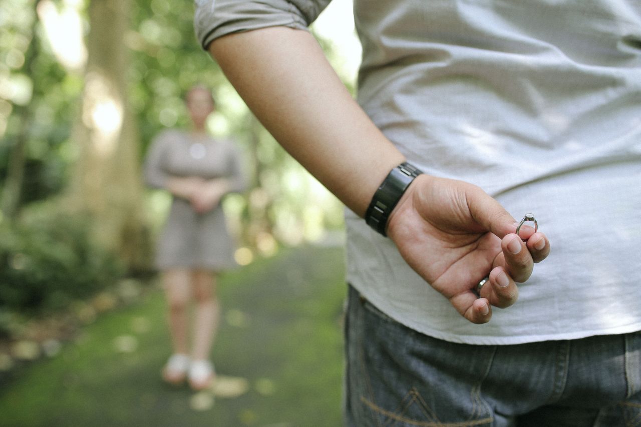 A man holds an engagement ring behind his back while a woman walks towards him.