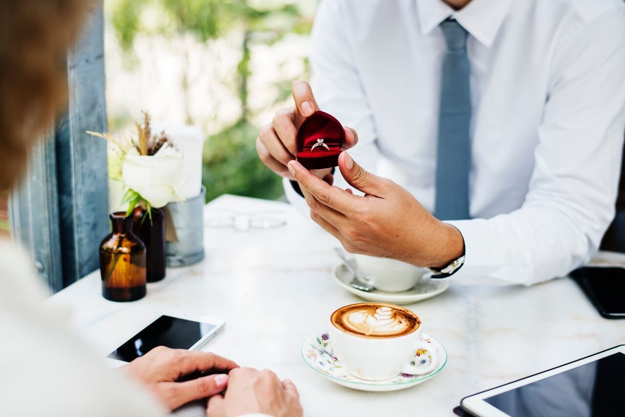 A man proposes to a woman with a diamond ring inside a red box, while sitting at a table with two coffees on it.