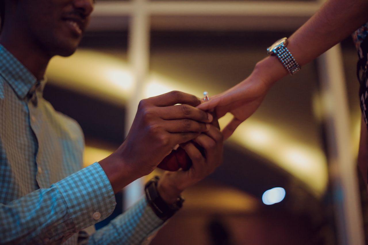 A man in a checkered shirt puts a ring on a woman&rsquo;s hand in front of warm lighting.