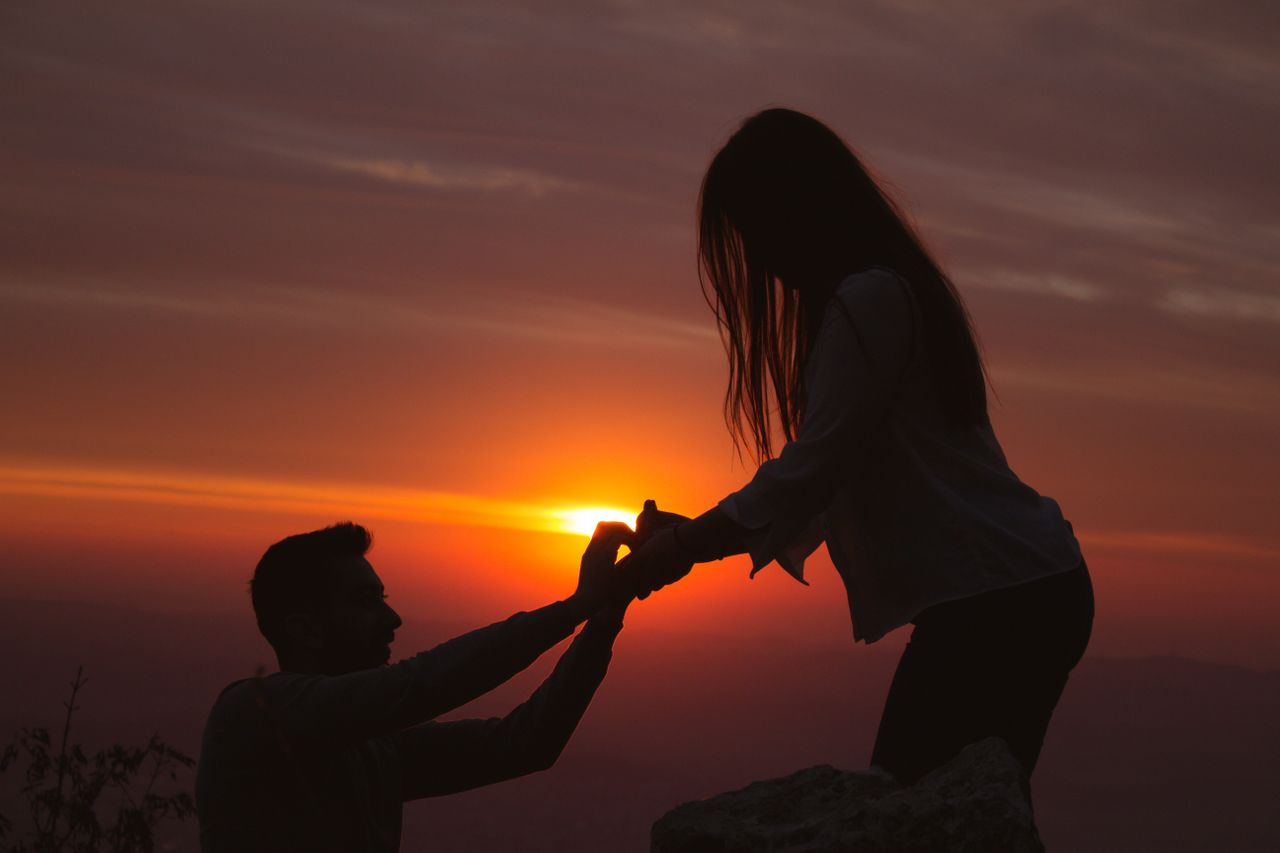 A couple getting engaged in front of the sunset, the man down on one knee with their silhouettes backlit.