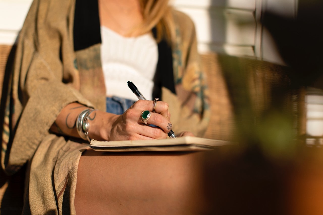 A woman writing in a notebook she’s balancing in her lap, her hand adorned with a gorgeous ring with an emerald gemstone and a silver bracelet.