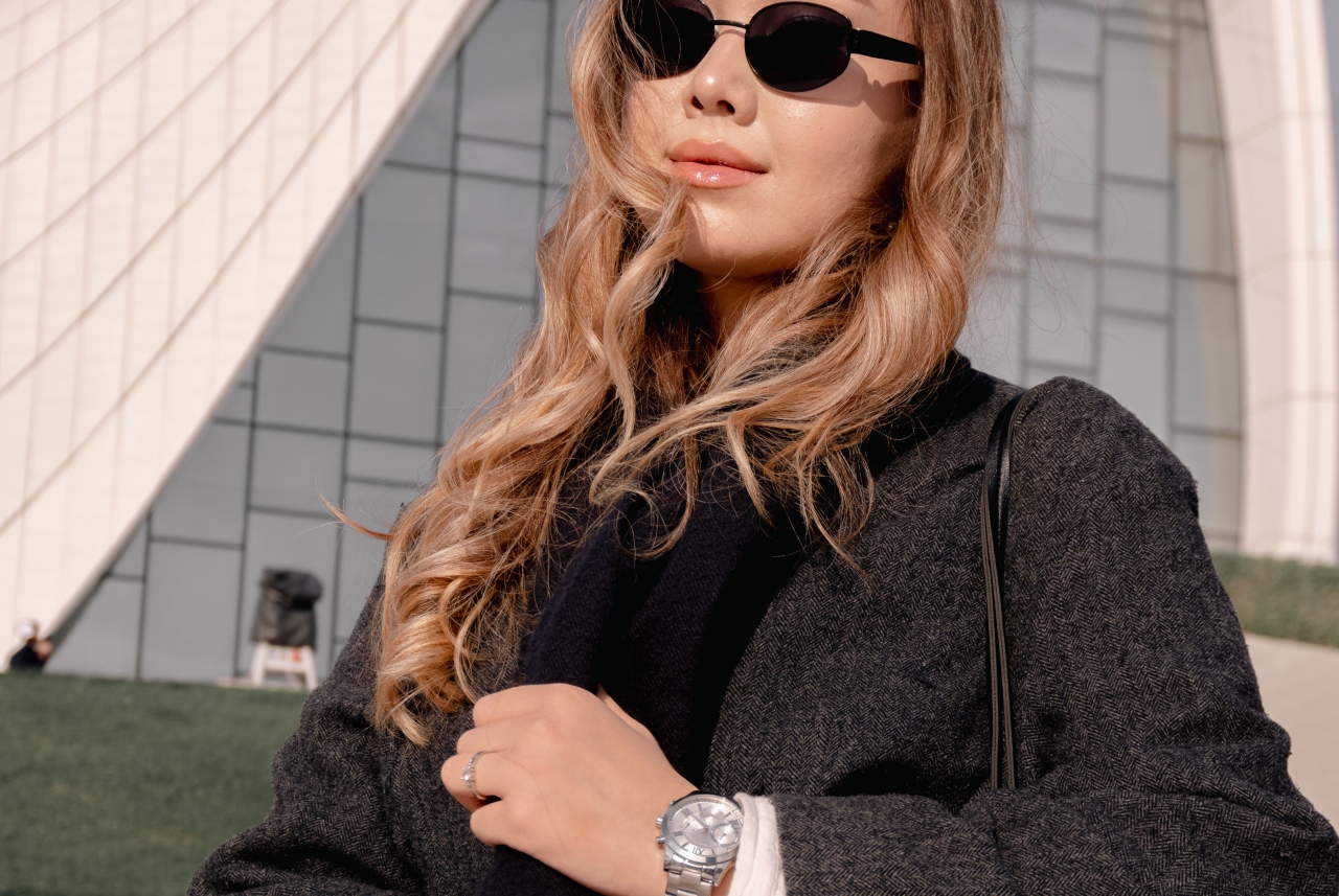 A close-up of a woman with blonde wavy hair wearing sunglasses, a dark coat, and a silver watch against a modern geometric building backdrop.