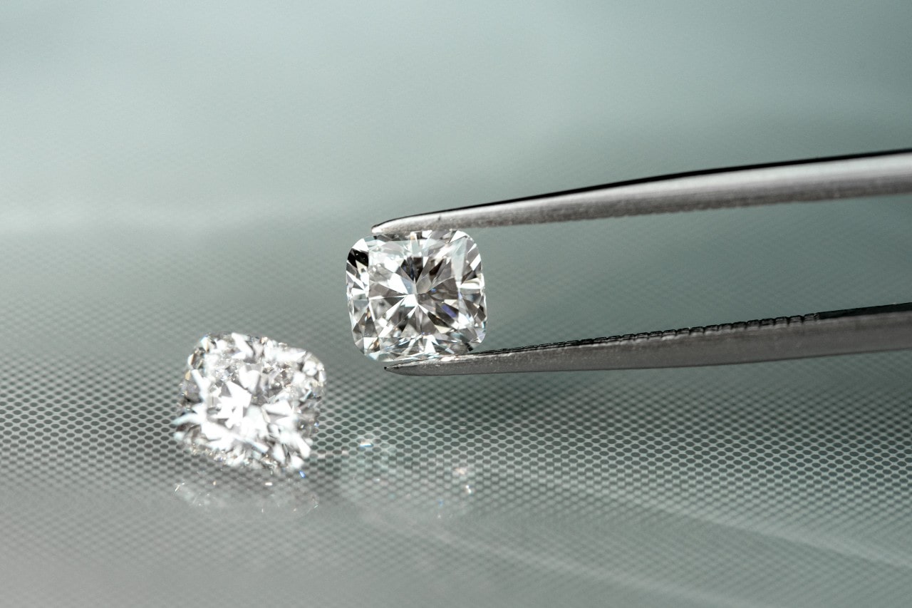 A close-up of a diamond being held by a pair of pliers, with another diamond resting next to it on the table.