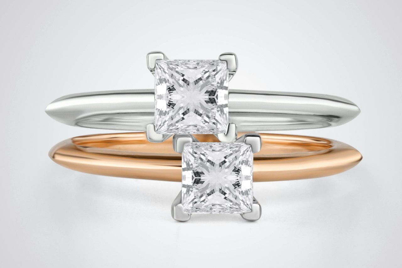 A silver engagement ring and a gold engagement ring stacked on top of one another, with a white background.