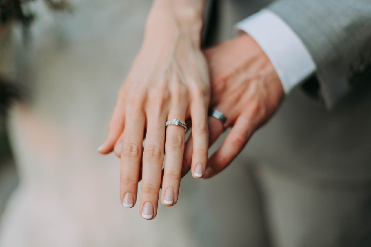 A close-up of a bride and groom's hands showing their wedding rings, with the bride wearing a diamond engagement ring and French manicure.