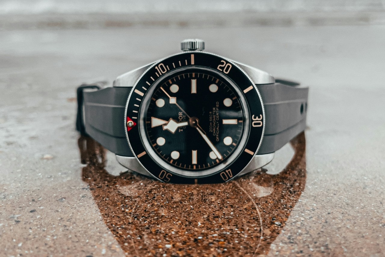 A TUDOR watch with a rubber strap and a black dial, set on sand.