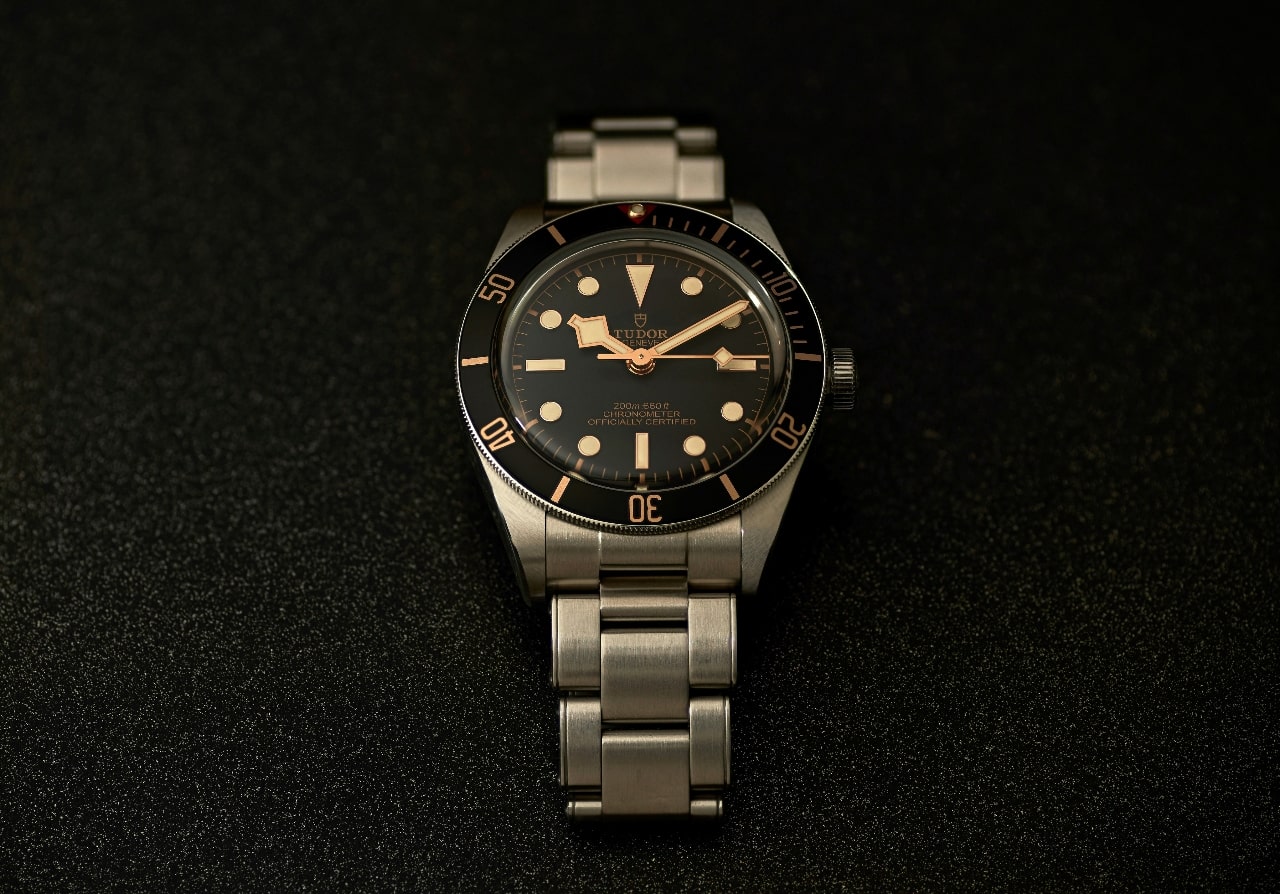 A TUDOR metallic watch with black dial on a black textured surface.