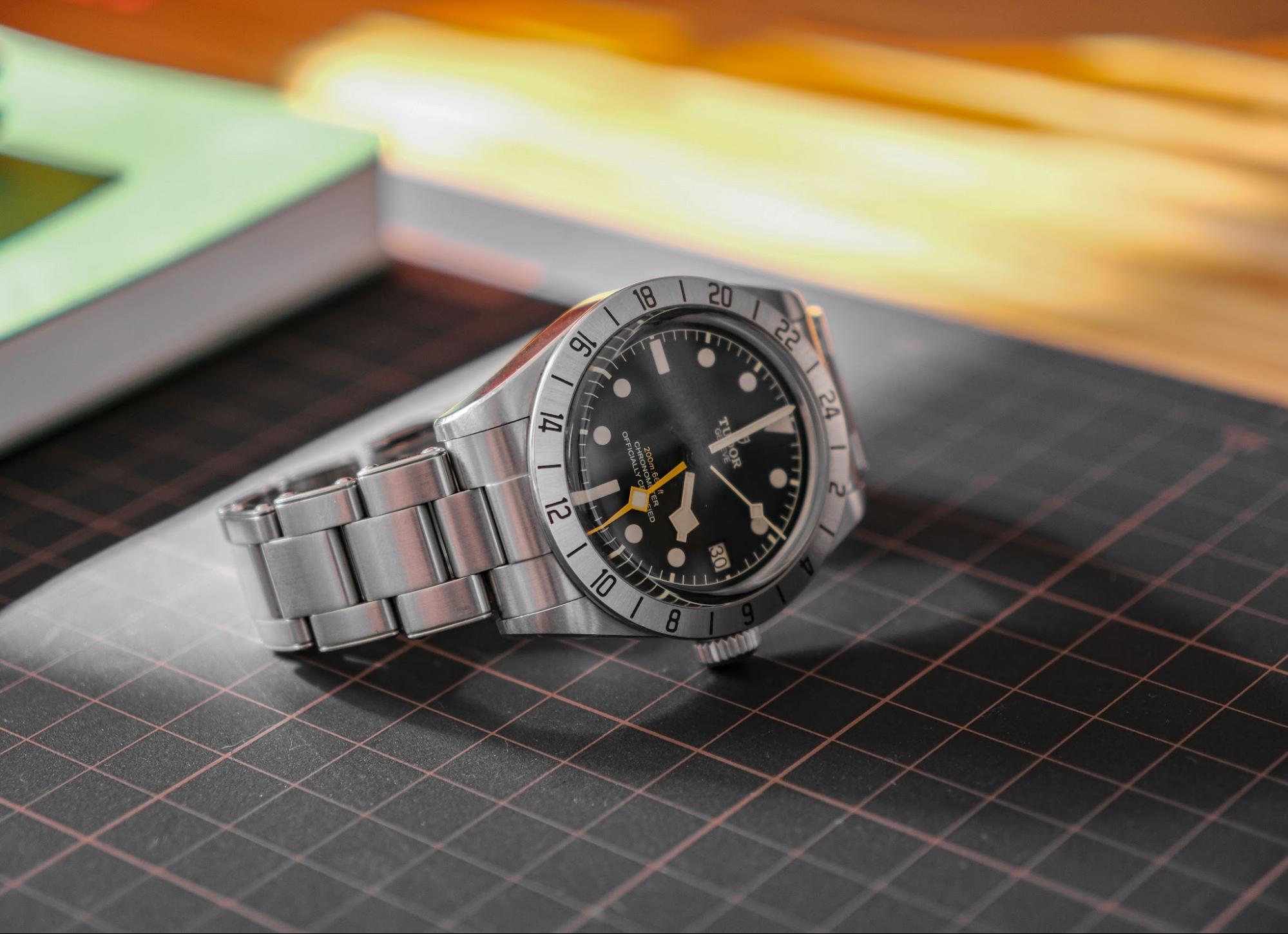 A flat-lay view of a stainless steel TUDOR watch resting on a black grid mat next to a notebook.