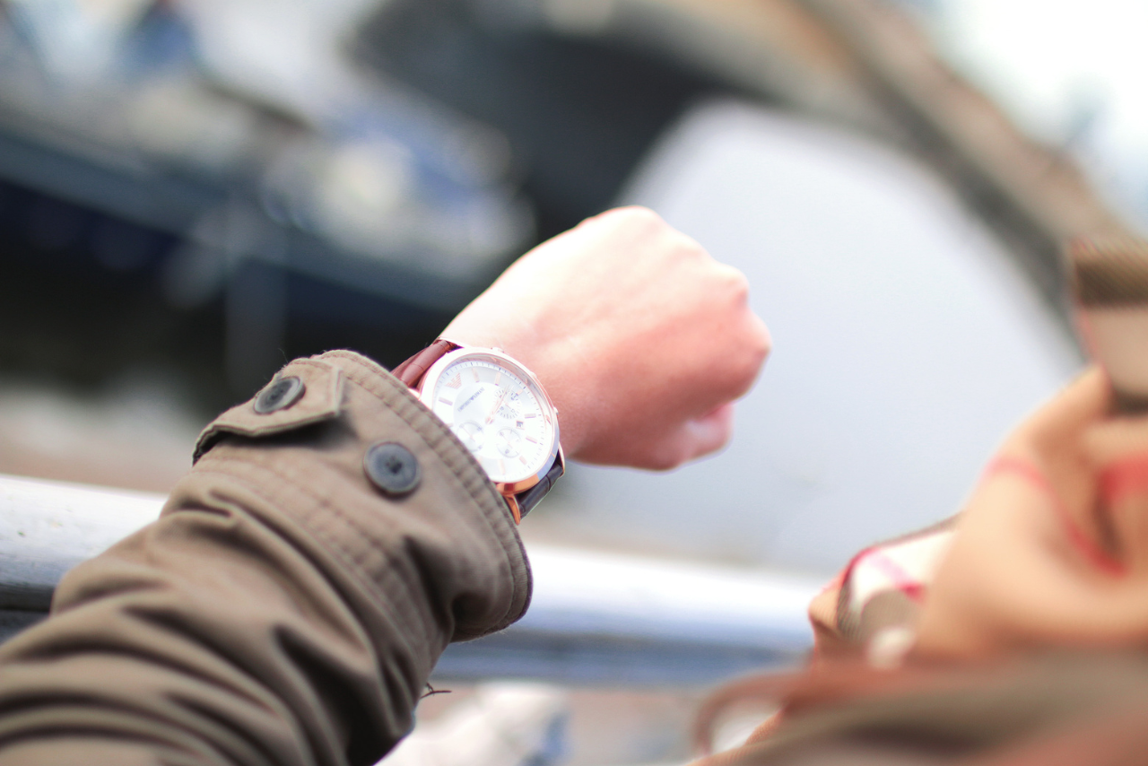 A close-up of a person wearing a brown trench coat while checking their luxury silver wristwatch.