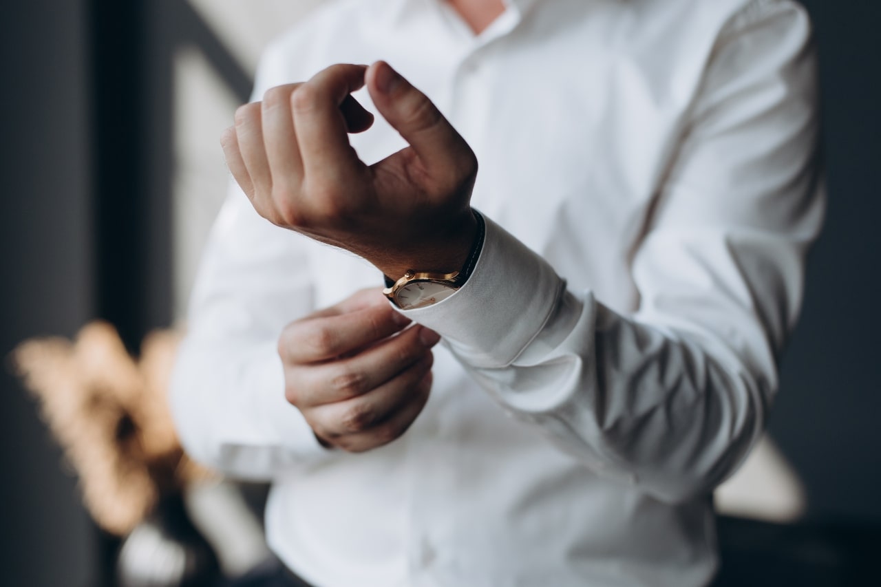 A man in a white dress shirt getting dressed for the day, adjusting his shirt cuffs and showcasing his watch.