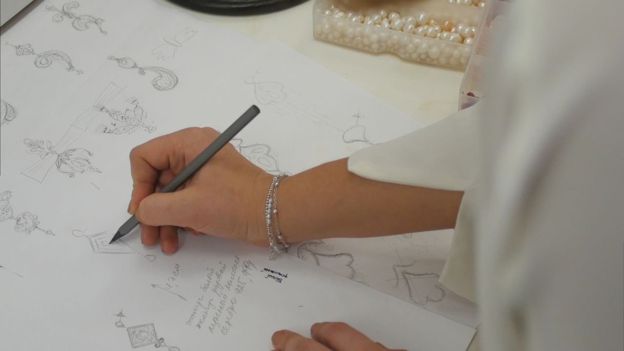 A jeweler drawing jewelry designs on a large white surface with a box of pearls in the background.