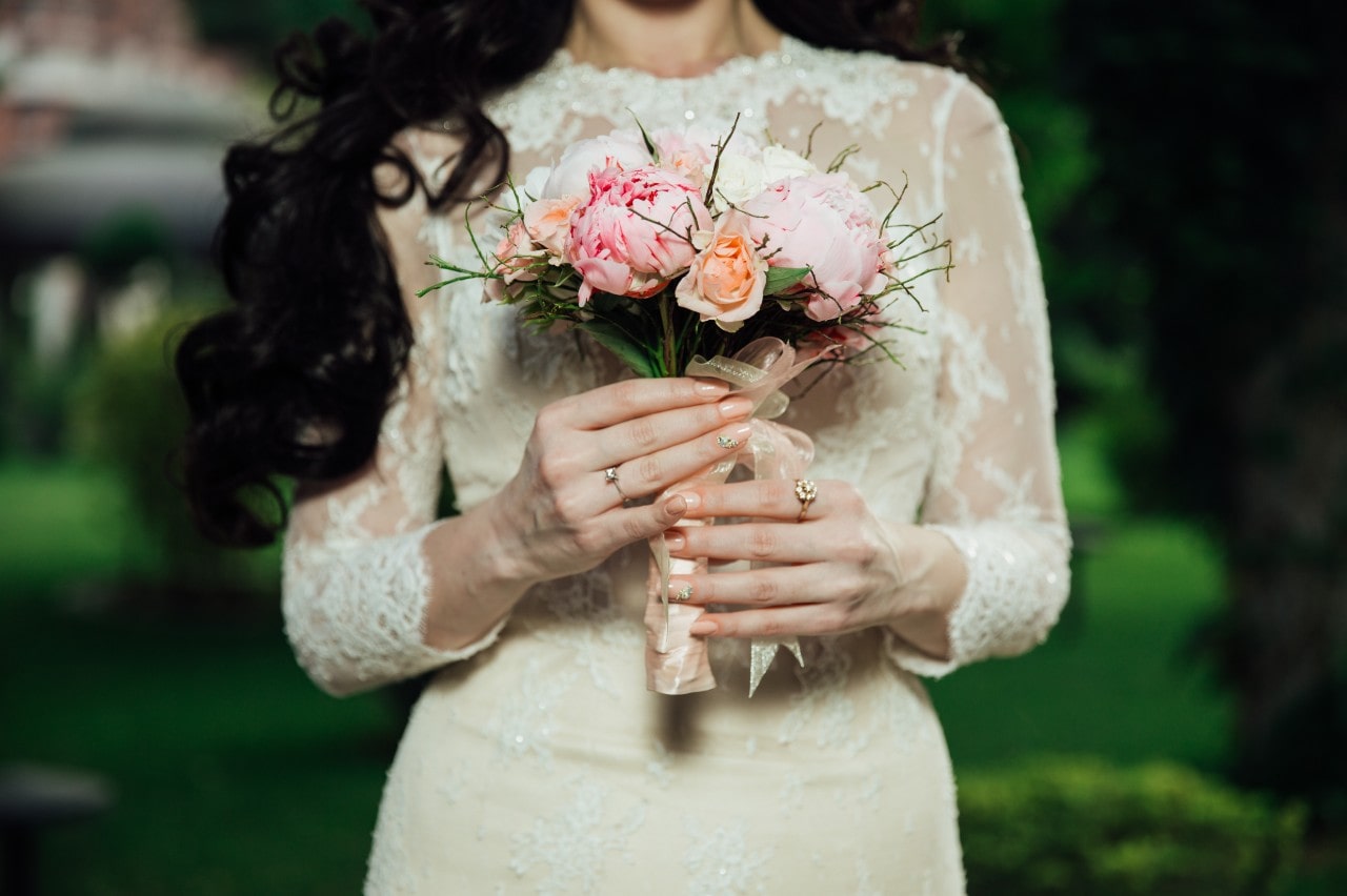 A close up of a bride in a lace wedding gown holding a bouquet of flowers, showcasing yellow gold engagement ring