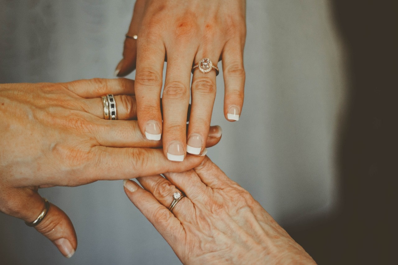Three women's hands of different ages held together, showcasing three generations of engagement rings and wedding bands.