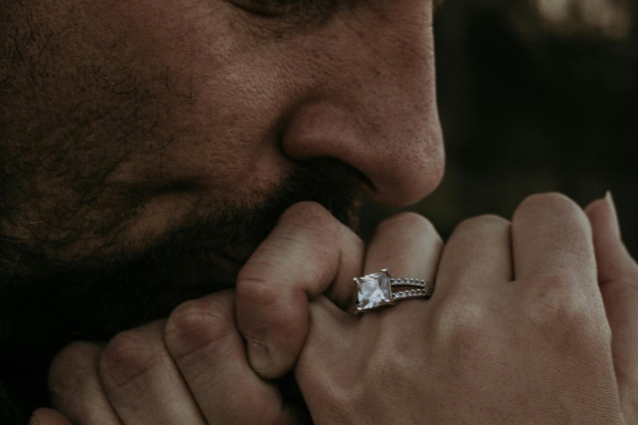 A man kissing a woman’s hand that is adorned with a princess cut engagement ring with side stones.