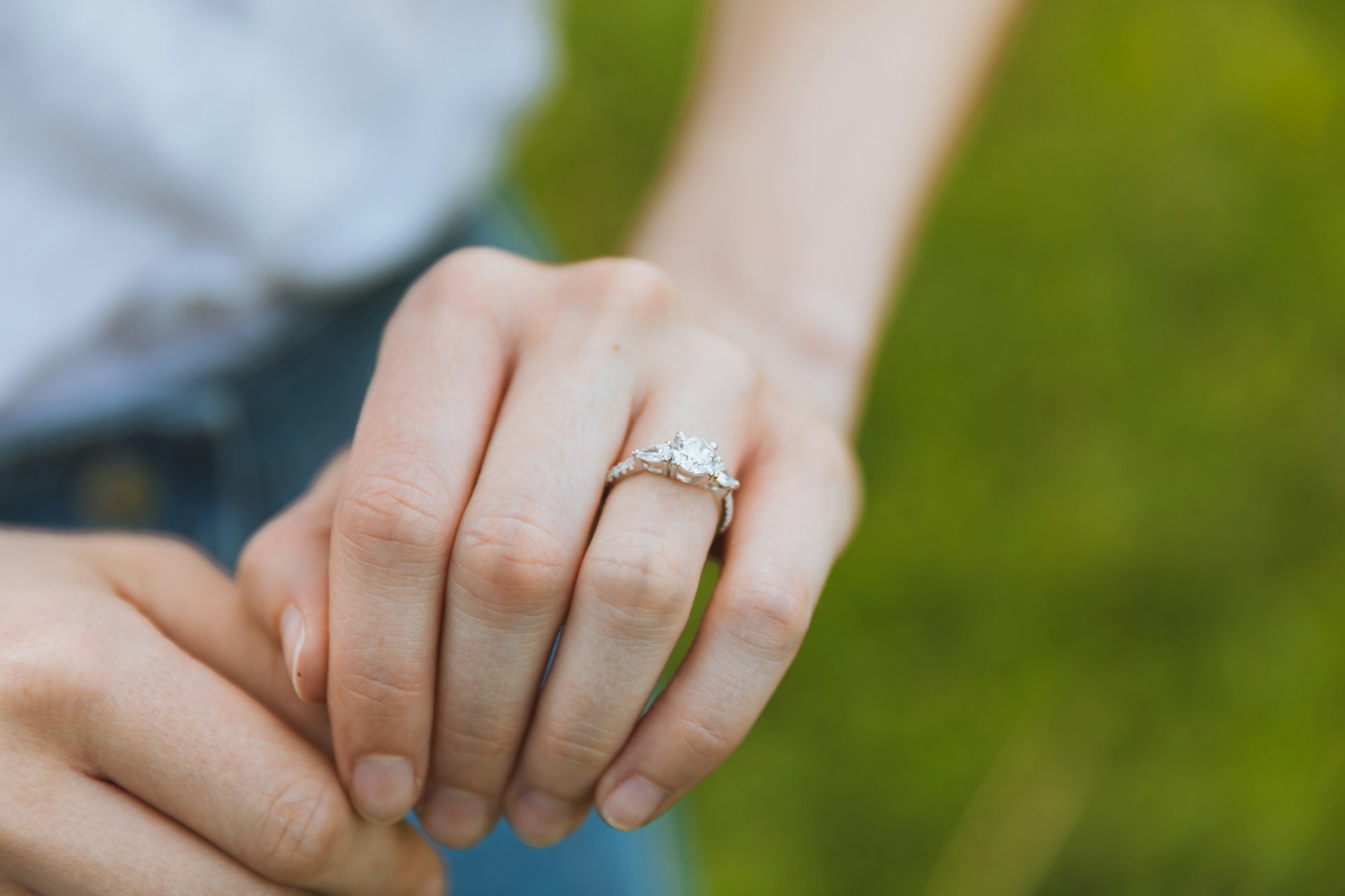 A close-up of a three stone white gold engagement ring displayed on a woman’s finger against a blurry grass