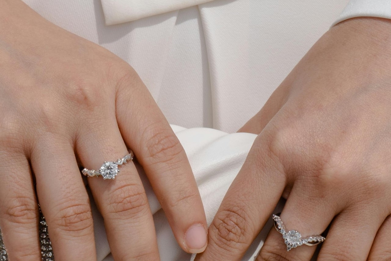 A close-up of two hands showcases elegant diamond engagement rings with intricate band designs against a soft white fabric background.