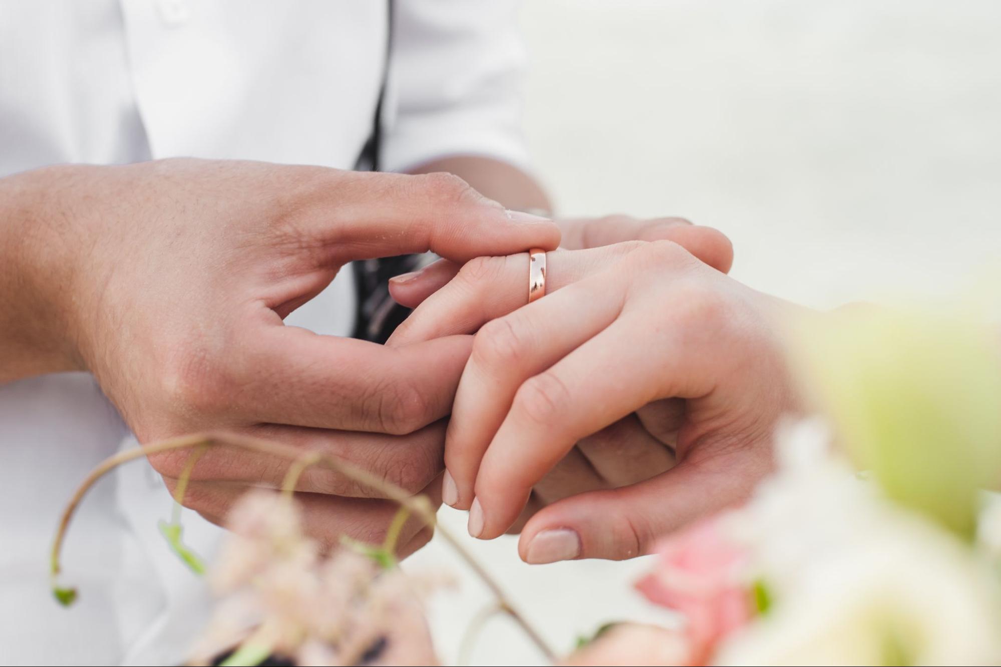A close-up of a man putting a yellow gold band on a woman’s hand with the blurred flowers in the front