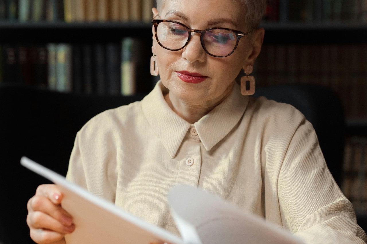 A lady in beige shirt and glasses reading a magazine, while sitting in a library, and showcasing geometric drop earrings.