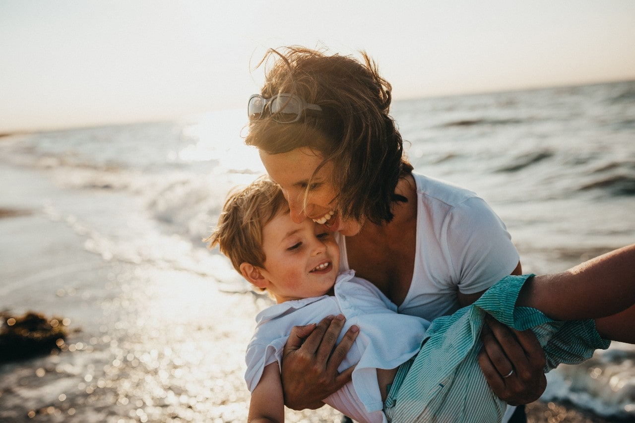 A woman embraces her son at the beach, both smiling as the sunlight reflects off the ocean waves.