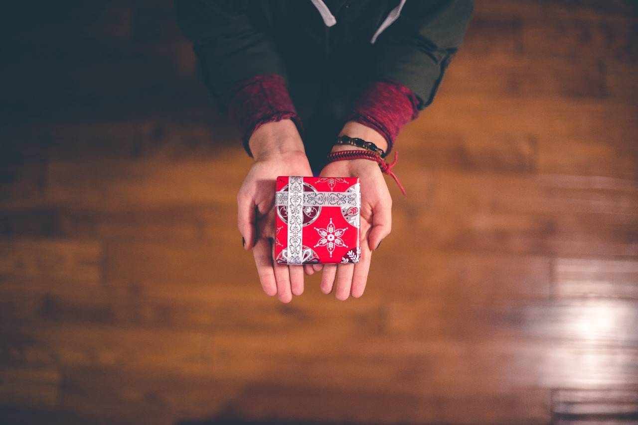 Hands holding a small, red gift box with intricate silver ribbon against a wooden floor background.